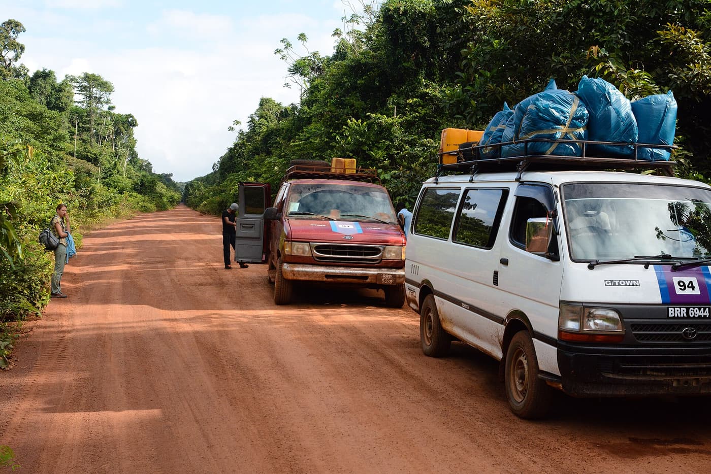 Two minibuses with luggage on their roofs stopped on a red dirt road through the forest