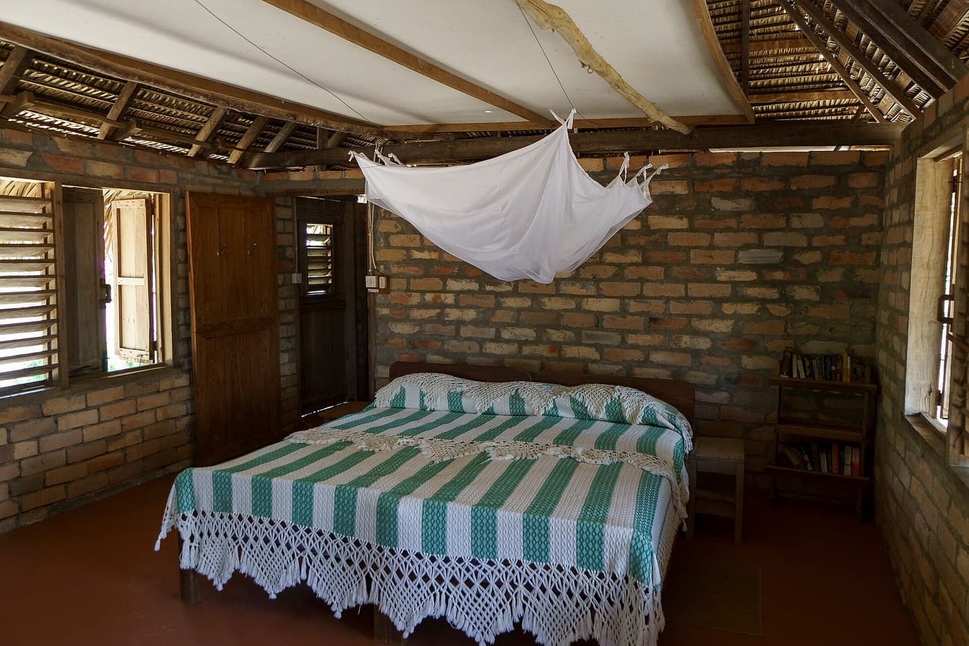A simple lodge bedroom with brick walls, a thatched roof and a mosquito net over the bed